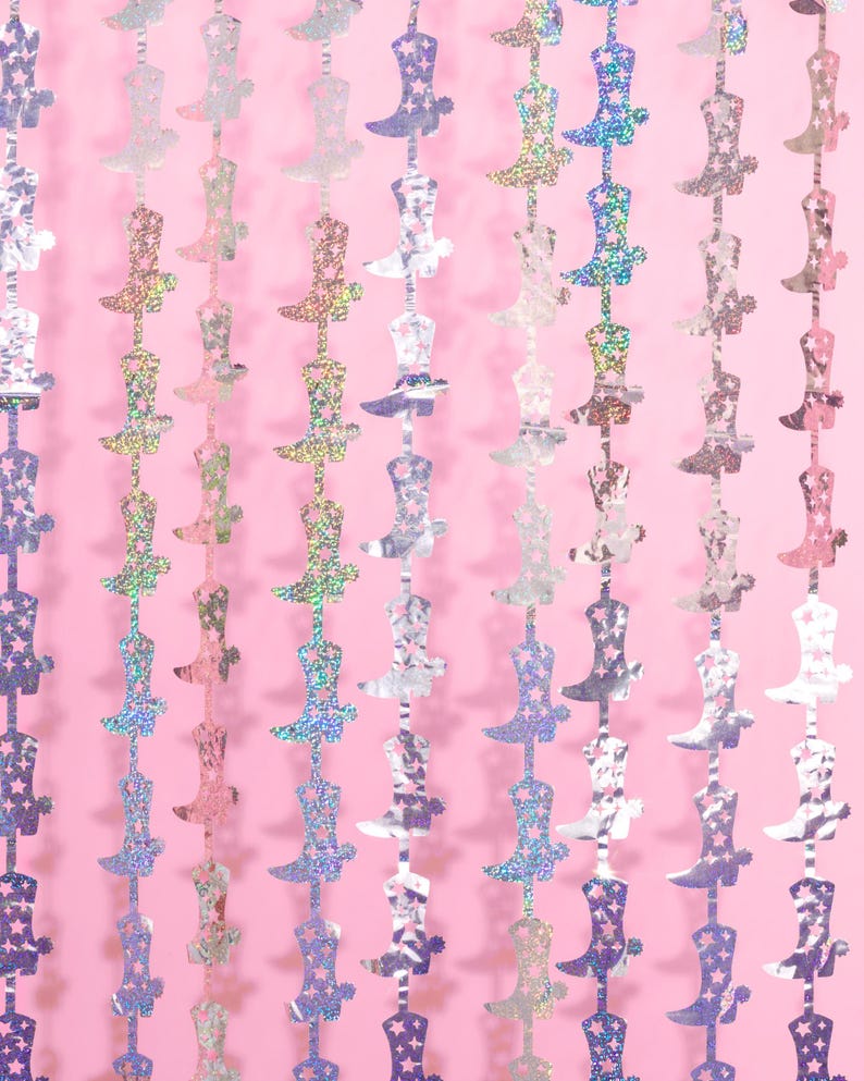 May include: A silver and iridescent garland of cowboy boots with stars. The garland is hanging against a pink background.