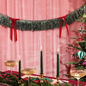 Puede incluir: Una escena festiva navide&ntilde;a con un &aacute;rbol de Navidad decorado, velas encendidas y copas de champ&aacute;n. Una guirnalda de oropel verde con lazos rojos cuelga sobre la mesa. El fondo es rosa.