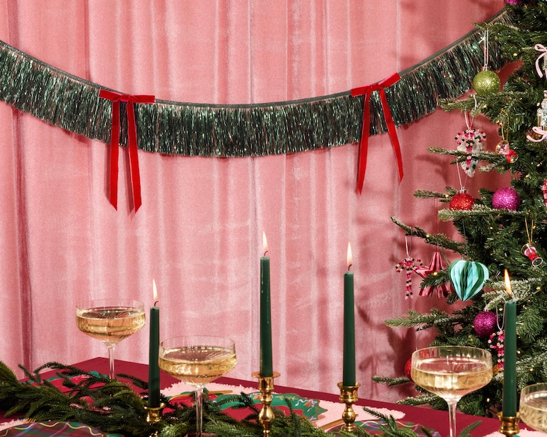 Puede incluir: Una escena festiva navide&ntilde;a con un &aacute;rbol de Navidad decorado, velas encendidas y copas de champ&aacute;n. Una guirnalda de oropel verde con lazos rojos cuelga sobre la mesa. El fondo es rosa.