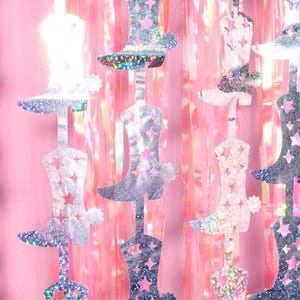 May include: A silver glitter cowboy boot garland with pink iridescent foil streamers. The garland is made of multiple silver glitter cowboy boots connected together with string.