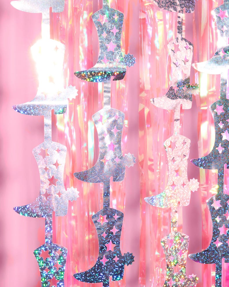 May include: A silver glitter cowboy boot garland with pink iridescent foil streamers. The garland is made of multiple silver glitter cowboy boots connected together with string.