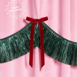 Puede incluir: Primer plano de una tela de terciopelo rosa con un lazo de terciopelo rojo oscuro y largas cintas. El lazo est&aacute; centrado sobre un fleco verde oscuro. El texto en forma de estrella blanca dice "VELVET RED BOWS".