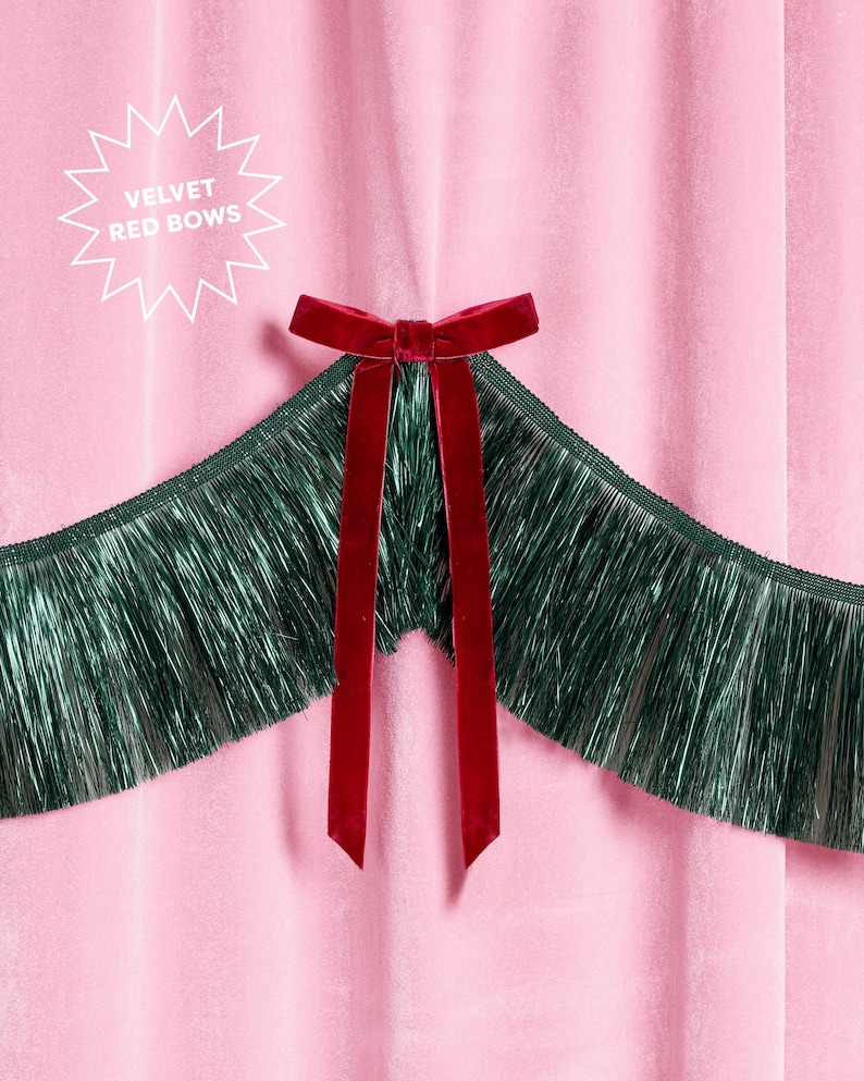 Puede incluir: Primer plano de una tela de terciopelo rosa con un lazo de terciopelo rojo oscuro y largas cintas. El lazo est&aacute; centrado sobre un fleco verde oscuro. El texto en forma de estrella blanca dice "VELVET RED BOWS".