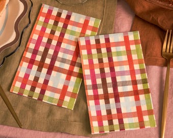May include: Two rectangular paper napkins with a colorful plaid pattern. The plaid design features a grid of red, pink, orange, green, and blue lines. The napkins are placed on a green and pink surface, with a gold fork and plate visible.