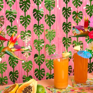 May include: Two tall glasses filled with orange juice, each topped with a small paper umbrella and fruit slice. The scene includes tropical flowers in glass vases, a halved papaya, a lime, and a backdrop of hanging green monstera leaf garlands.
