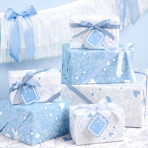 May include: A collection of wrapped presents in blue and white, adorned with ribbons and tags. The wrapping paper displays a pattern of hearts and other designs. The gifts are arranged on a patterned surface, with a blue backdrop.