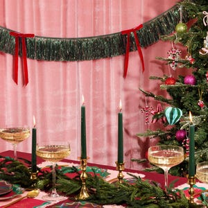 Puede incluir: Una mesa festiva navide&ntilde;a con un mantel rojo, guirnalda verde y candelabros dorados con velas verdes encendidas. Copas de champ&aacute;n y platos a cuadros est&aacute;n listos para una celebraci&oacute;n. Un &aacute;rbol de Navidad y una guirnalda de oropel verde con lazos rojos completan la escena.
