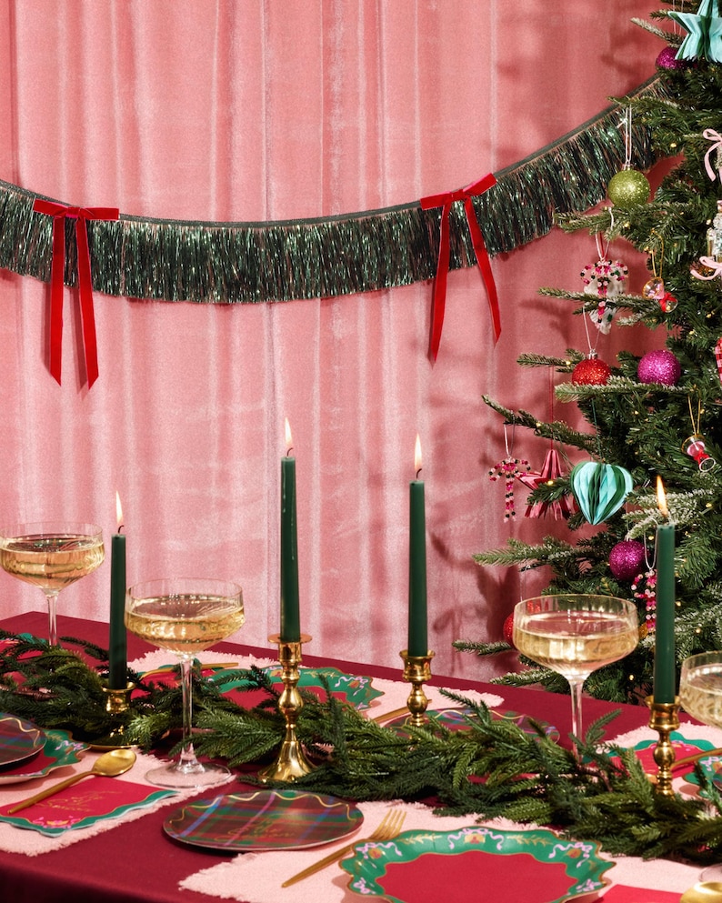 Puede incluir: Una mesa festiva navide&ntilde;a con un mantel rojo, guirnalda verde y candelabros dorados con velas verdes encendidas. Copas de champ&aacute;n y platos a cuadros est&aacute;n listos para una celebraci&oacute;n. Un &aacute;rbol de Navidad y una guirnalda de oropel verde con lazos rojos completan la escena.