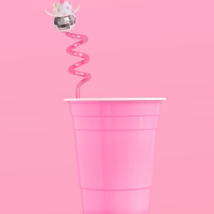 May include: A pink plastic cup with a white rim sits on an orange cylinder against a pink background. A pink swirly straw extends from the cup, topped with a disco ball wearing a white cowboy hat.