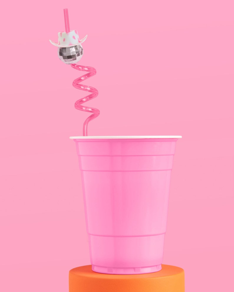May include: A pink plastic cup with a white rim sits on an orange cylinder against a pink background. A pink swirly straw extends from the cup, topped with a disco ball wearing a white cowboy hat.