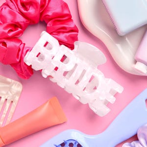 May include: A collection of beauty accessories on a pink background. Includes a white hair clip with the word "BRIDE", a pink satin scrunchie, a blue hairbrush, and tubes of lip gloss. The items are arranged in a flat lay style.