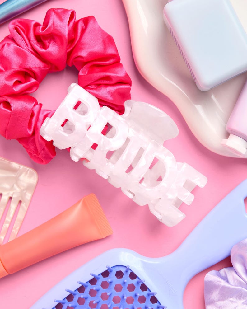 May include: A collection of beauty accessories on a pink background. Includes a white hair clip with the word "BRIDE", a pink satin scrunchie, a blue hairbrush, and tubes of lip gloss. The items are arranged in a flat lay style.