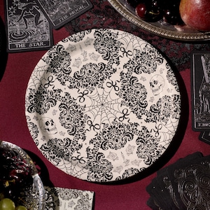 May include: A round, off-white plate with a black damask pattern, spiderwebs, and skull accents. The plate is surrounded by tarot cards and a silver serving tray with fruit. The overall theme is gothic and Halloween.