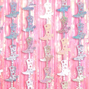 May include: A garland of iridescent silver and pink cowboy boots with stars on them. The boots are hanging on a pink and iridescent background.