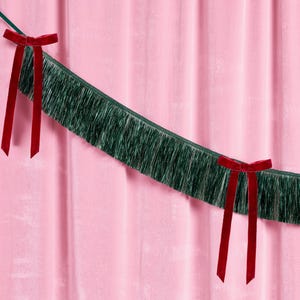 Puede incluir: Un fondo de cortina de terciopelo rosa con una guirnalda de flecos verdes y dos lazos de terciopelo rojo. La guirnalda est&aacute; tendida a trav&eacute;s de la imagen, y los lazos est&aacute;n atados a la guirnalda.