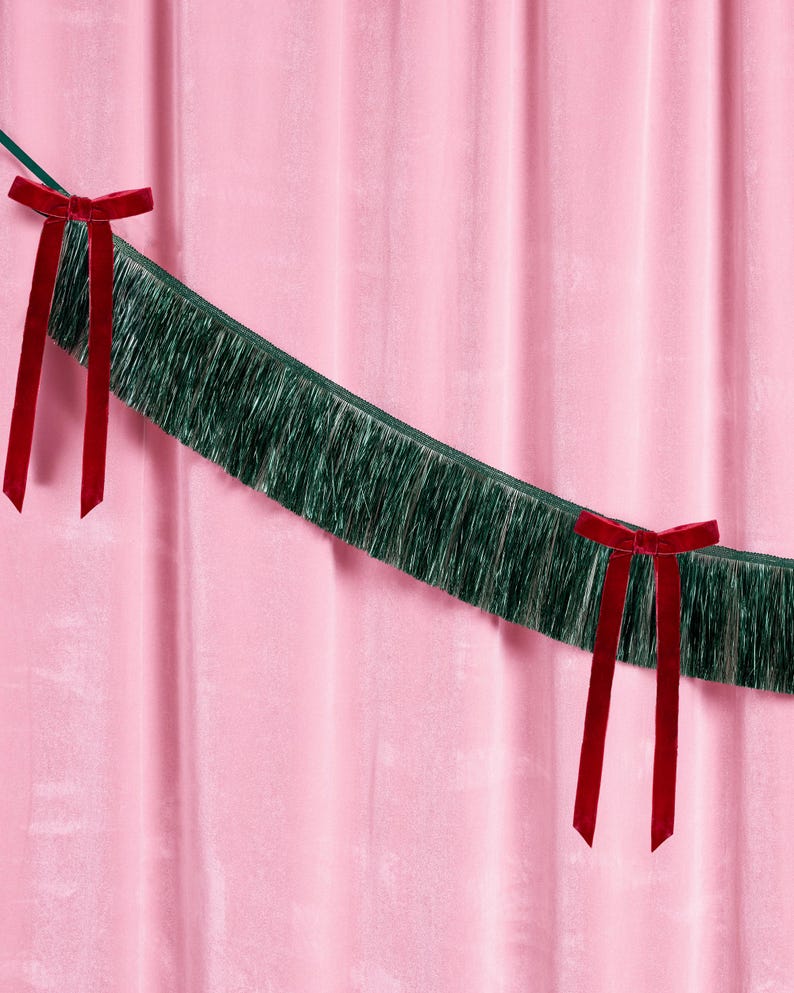 Puede incluir: Un fondo de cortina de terciopelo rosa con una guirnalda de flecos verdes y dos lazos de terciopelo rojo. La guirnalda est&aacute; tendida a trav&eacute;s de la imagen, y los lazos est&aacute;n atados a la guirnalda.