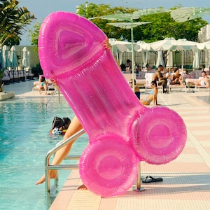 May include: A pink inflatable pool float shaped like a human body part. The float is in a swimming pool with a person standing on the edge of the pool.