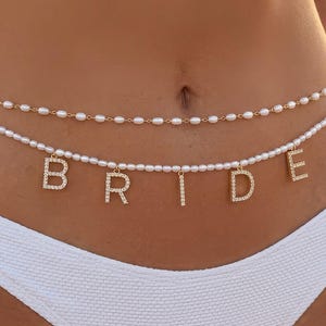 May include: A two-strand waist chain with pearls and gold accents. The lower strand spells out the word "BRIDE" with gold-colored, rhinestone-studded letters. The chain is worn over a white bikini bottom.