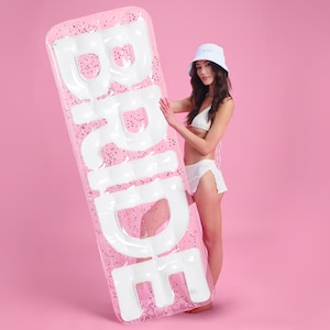 May include: Pink inflatable pool float with the word "Bride" in white letters. A person is holding the float in front of a pink background.