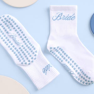 May include: White socks with the word "Bride" embroidered in light blue. The soles feature a pattern of small, light blue hearts for grip. The socks are on a light blue background.