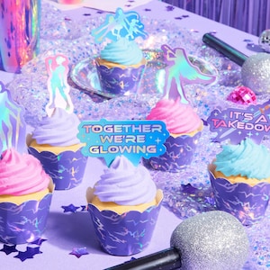 May include: A collection of cupcakes with pastel frosting in purple cupcake wrappers. Each cupcake is decorated with a holographic figure or a sign that reads "Together We're Glowing" or "It's a Takedown". The scene is set on a purple surface with scattered confetti.