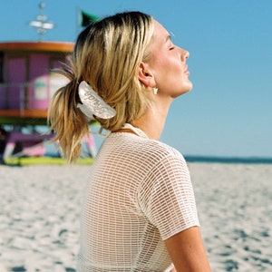 K&ouml;nnte beinhalten: Eine Frau mit blonden Haaren tr&auml;gt ein wei&szlig;es Netz-Crop-Top mit einem wei&szlig;en Haarklipp. Sie sitzt an einem Sandstrand mit einem Rettungsschwimmerstand im Hintergrund.