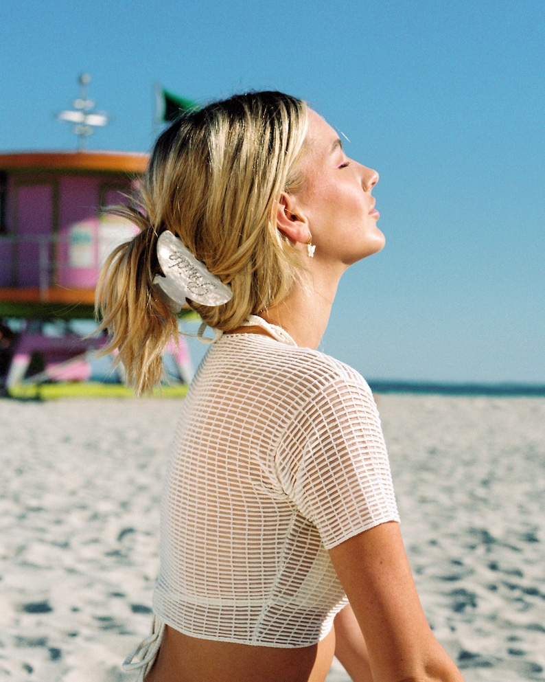 K&ouml;nnte beinhalten: Eine Frau mit blonden Haaren tr&auml;gt ein wei&szlig;es Netz-Crop-Top mit einem wei&szlig;en Haarklipp. Sie sitzt an einem Sandstrand mit einem Rettungsschwimmerstand im Hintergrund.