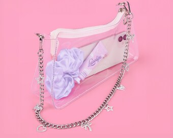 Puede incluir: Una bolsa de plástico transparente con una correa de cadena plateada. La bolsa contiene un tubo de bálsamo labial morado con el texto "Lanolips", una scrunchie blanca y una funda de teléfono blanca. La cadena tiene dijes de estrellas y corazones.