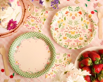 May include: A festive picnic scene with floral-patterned plates, napkins, and wooden forks. One plate has a green gingham border. A cake with flowers and a bowl of strawberries are also present, creating a cheerful, colorful display.