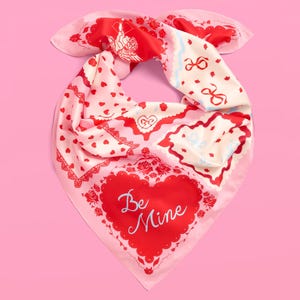 May include: A pink silk scarf with a Valentine's Day theme. The scarf features red hearts, roses, and the words "Be Mine" in a heart-shaped design. Other patterns include bows and cherubs.