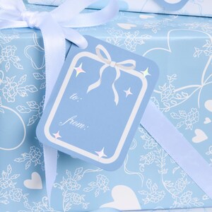 May include: A light blue gift box wrapped in patterned paper with white floral and heart designs. A matching gift tag with a bow and the words "to:" and "from:" is attached with a white ribbon. The tag has star accents.