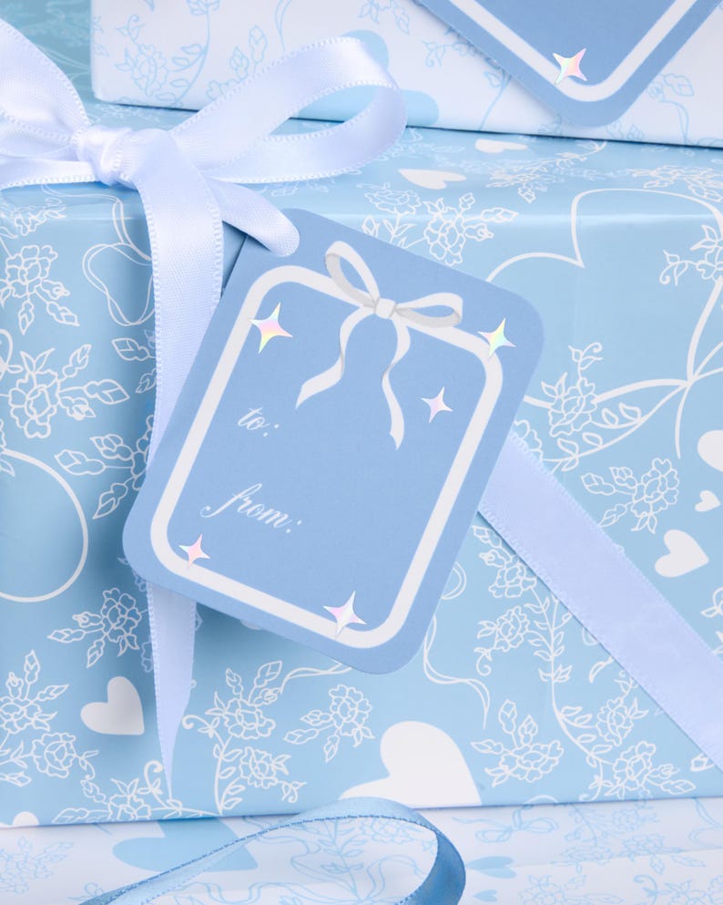 May include: A light blue gift box wrapped in patterned paper with white floral and heart designs. A matching gift tag with a bow and the words "to:" and "from:" is attached with a white ribbon. The tag has star accents.