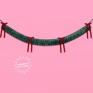Puede incluir: Una guirnalda de oropel verde con lazos rojos, sobre un fondo rosa. La guirnalda mide 1,83 metros de largo, como indica el texto en un gr&aacute;fico de estrella blanca. El oropel tiene una textura de flecos.