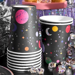 May include: Black paper cups with a space-themed design. The cups feature colorful planets, stars, and moons. A stack of cups is visible, along with a single cup. The cups are decorated with silver foil accents and have white rims.