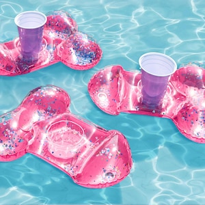 May include: Three pink inflatable drink holders with a clear cup holder and a purple cup in each. The holders are shaped like a figure eight and are covered in glitter. They are floating in a pool of blue water.
