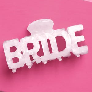 May include: A white, marbled-effect hair claw clip spelling out the word "BRIDE". The clip is set against a bright pink background. This accessory is perfect for a bridal shower or wedding.