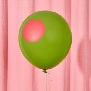 May include: A green balloon with a pink circle on its surface. The balloon is tied with a silver string and set against a pink backdrop. The balloon's shape is round and inflated.
