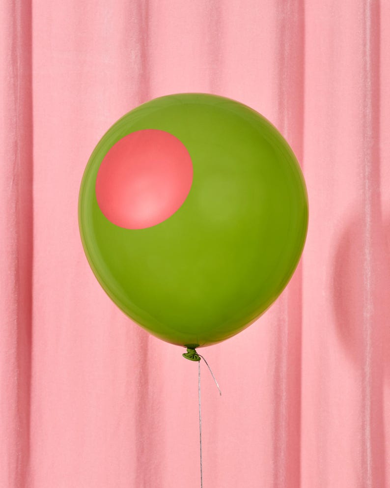 May include: A green balloon with a pink circle on its surface. The balloon is tied with a silver string and set against a pink backdrop. The balloon's shape is round and inflated.