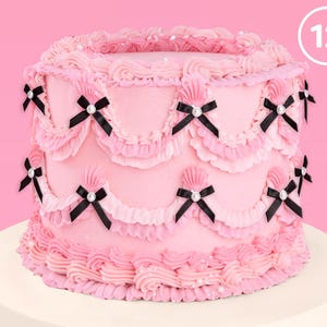 May include: A two-tiered pink cake decorated with black and white bows, pink frosting, and white pearl sprinkles. The cake is on a white cake stand. The number 12 is written in white on a pink circle.