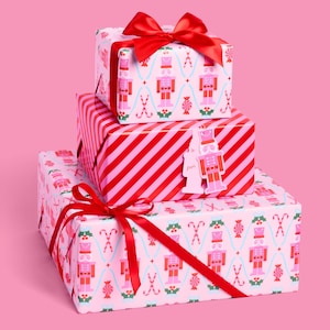 May include: Three wrapped gifts stacked on top of each other. The top and bottom gifts are wrapped in light pink paper with a pattern of nutcrackers, candy canes, and holly. The middle gift is wrapped in red and pink diagonal stripes. Each gift is tied with a red ribbon.