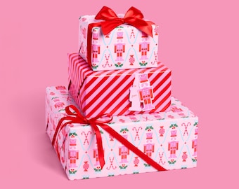 Könnte beinhalten: Drei gestapelte Weihnachtsgeschenke. Das obere und untere Geschenk sind in hellrosa Papier mit einem Muster aus Nussknackern, Zuckerstangen und Stechpalmen verpackt. Das mittlere Geschenk ist in rot-rosa diagonalen Streifen verpackt. Jedes Geschenk ist mit einem roten Band gebunden.