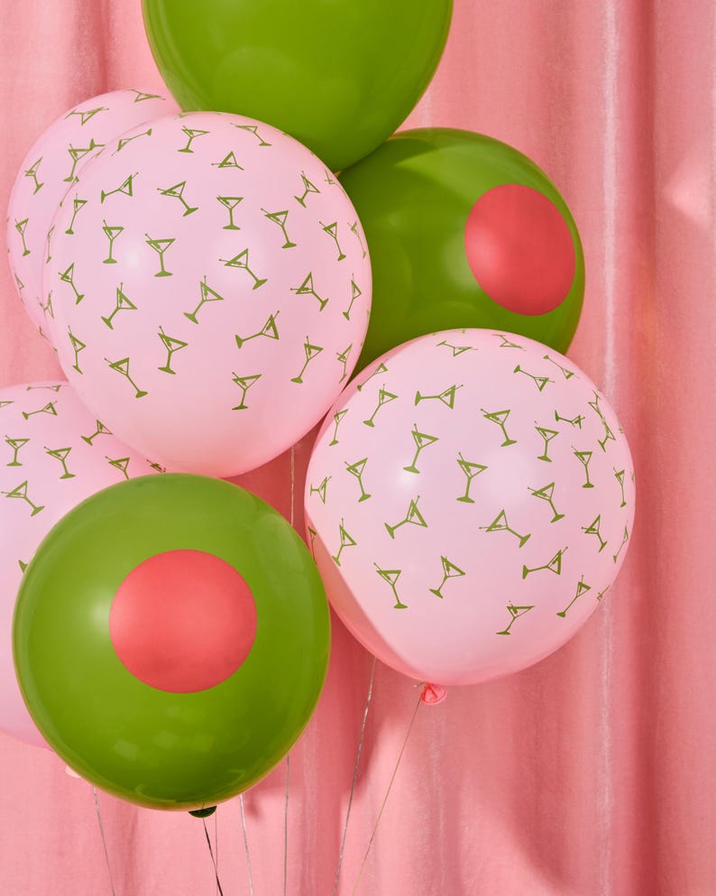 May include: A cluster of party balloons in shades of pink and green. Some pink balloons are patterned with small martini glass illustrations. Green balloons have a red circle design. The balloons are tied together with silver string.