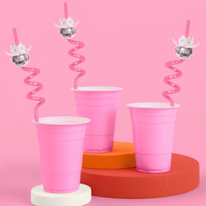 May include: Three pink plastic cups with white rims are displayed on a pink background. Each cup has a pink, curly straw and is topped with a disco ball wearing a white cowboy hat. The cups are on a white, orange, and red platform.