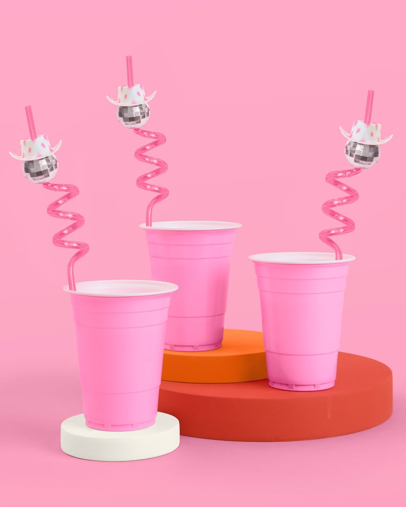 May include: Three pink plastic cups with white rims are displayed on a pink background. Each cup has a pink, curly straw and is topped with a disco ball wearing a white cowboy hat. The cups are on a white, orange, and red platform.