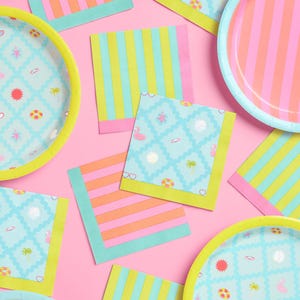 May include: A collection of colorful party supplies, including paper plates and napkins, arranged on a pink surface. The plates and napkins feature bright stripes and summer-themed patterns in shades of pink, blue, yellow, and orange.