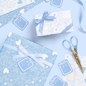 May include: Overhead view of gift wrapping supplies in blue and white. The scene includes wrapped gifts, gift tags, ribbon, and scissors. The wrapping paper features a pattern of hearts, bows, and floral designs. The gift tags have a bow design and the words "to from".