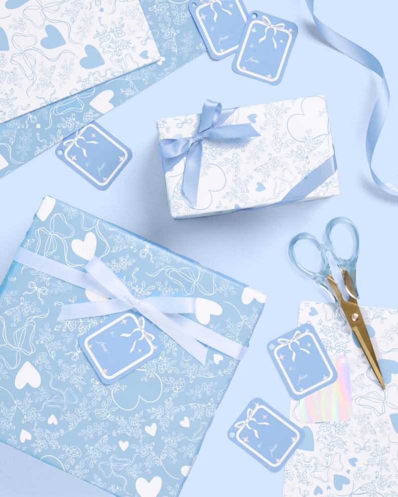 May include: Overhead view of gift wrapping supplies in blue and white. The scene includes wrapped gifts, gift tags, ribbon, and scissors. The wrapping paper features a pattern of hearts, bows, and floral designs. The gift tags have a bow design and the words "to from".
