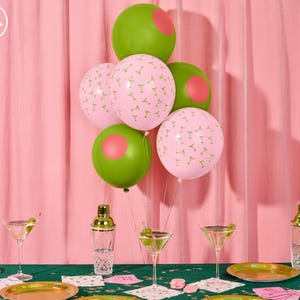 May include: A festive party scene with pink and green balloons, martini glasses, and gold-rimmed plates. The balloons are decorated with martini glass patterns and pink circles. The scene is set on a green tablecloth with a pink backdrop.