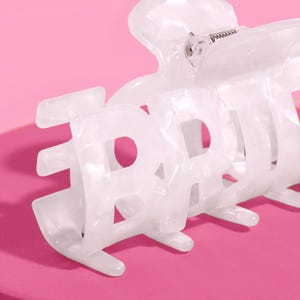 May include: White hair claw clip with the word "BABY" in a bold, open font. The clip has a marbled effect and a metal spring mechanism. The background is a solid pink.