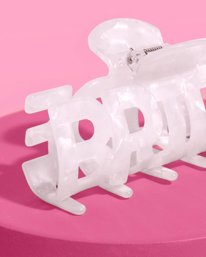 May include: White hair claw clip with the word "BABY" in a bold, open font. The clip has a marbled effect and a metal spring mechanism. The background is a solid pink.